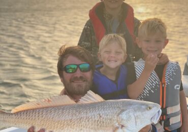 Photo of Captain Mark and his boys on a fishing boat holding a redfish.