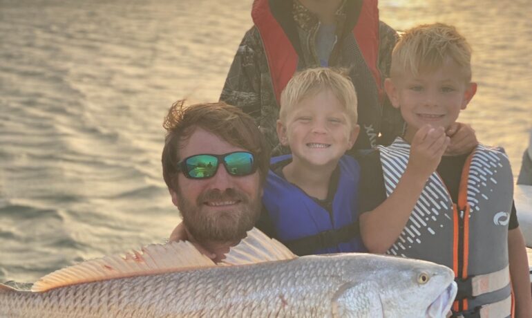 Photo of Captain Mark and his boys on a fishing boat holding a redfish.